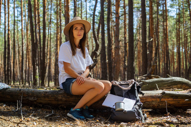 Young woman tourist in hat and t-shirt sits on a log during a halt in the forest. - Photo, Image
