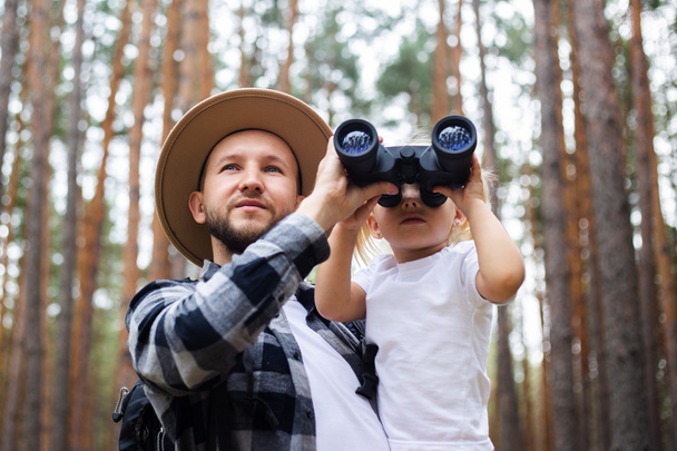Человек в шляпе, рюкзаке и ребенок смотрят в бинокль во время прогулки по лесу. Семейный поход в горы или лес. - Фото, изображение