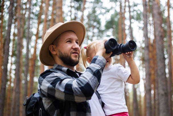 El hombre con un sombrero y una mochila y un niño miran a través de los prismáticos mientras caminan por el bosque. Caminata familiar a las montañas o al bosque. - Foto, Imagen