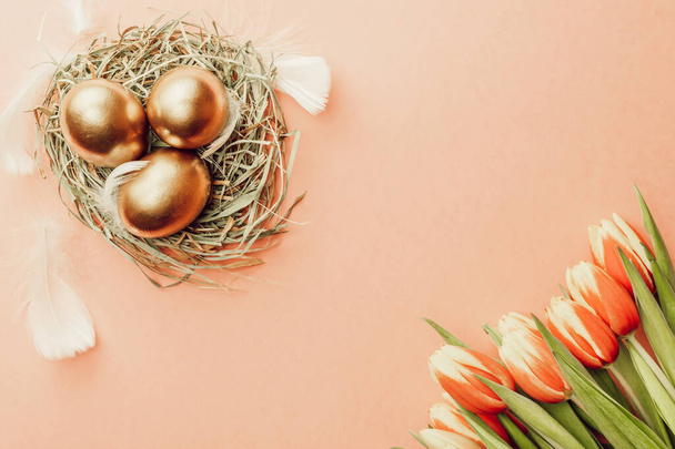 Golden easter colour eggs in basket with spring tulips, white feathers on pastel pink background in Happy Easter decoration. Spring holiday top view concept - Photo, Image