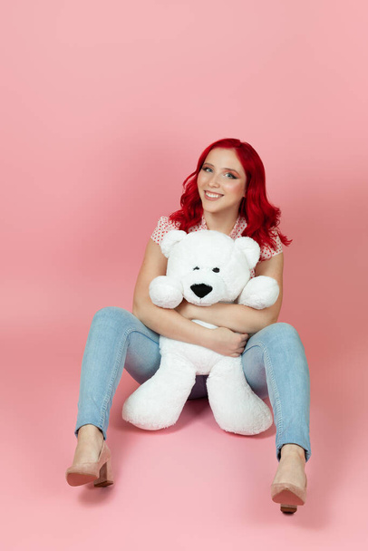 a full-length smiling, cheerful woman in jeans with red hair holds a large white teddy bear between her legs , isolated on a pink background - Fotó, kép
