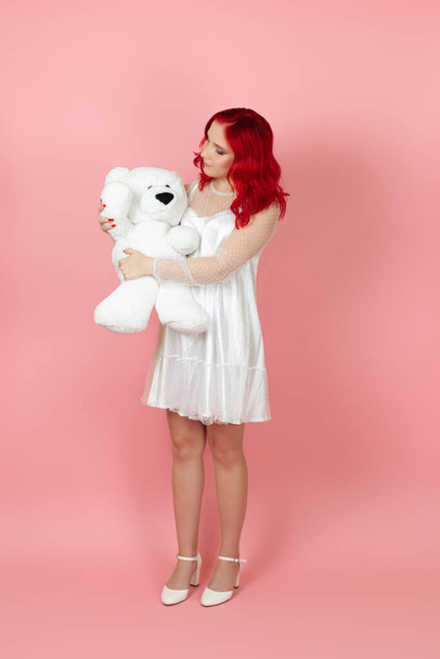 full-length charming woman in a white dress and with red hair holds a large white teddy bear isolated on a pink background - Photo, Image