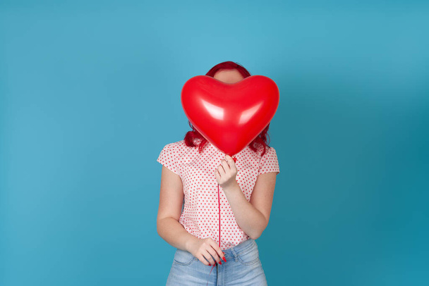 portrait of a woman with red hair hiding behind a red flying balloon in the form of a heart, isolated on a blue background - Photo, Image