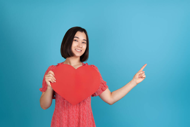 close-up a smiling, happy young Asian woman in a red dress holds a large red paper heart in her hands and points with her index finger at an empty space for text , isolated on a blue background - Foto, Bild