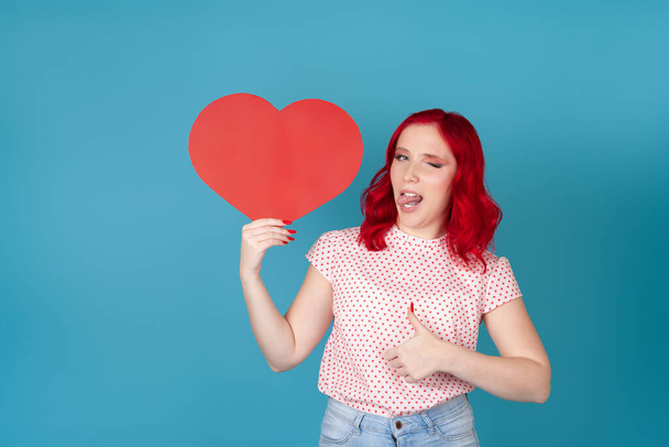 happy, joyful young woman with red hair holds red paper heart, winks, putting out tongue and gives thumbs up isolated on blue background - Zdjęcie, obraz
