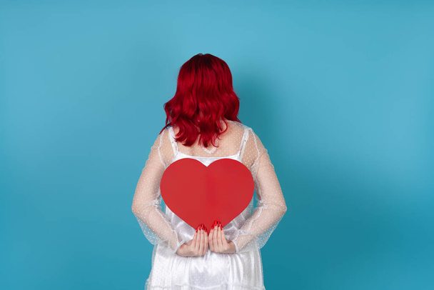 een jonge vrouw in een witte jurk en met rood haar houdt een groot rood papieren hart achter haar rug, geïsoleerd op een blauwe achtergrond - Foto, afbeelding