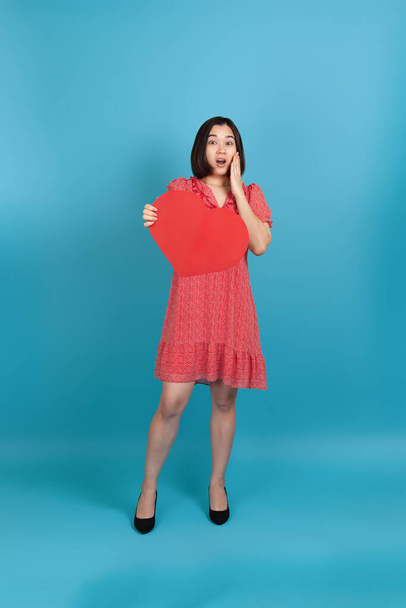 full-length shocked excited young asian woman with open mouth in red dress holding big red paper heart isolated on blue background - Фото, изображение