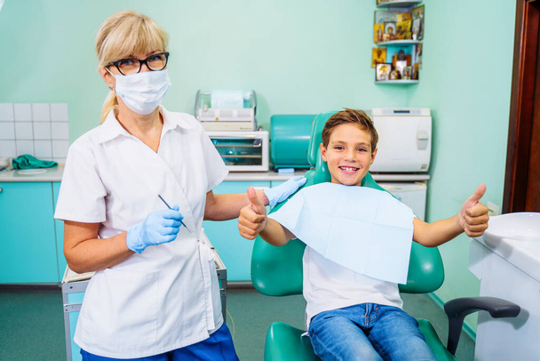 Der kleine, geduldige Junge freut sich über einen Zahnarztbesuch. Konzept der schmerzlosen Zahnbehandlung. Schönes, breites Lächeln eines Kindes, das in einem Zahnarztstuhl in einer Arztpraxis sitzt. Daumen hoch. - Foto, Bild