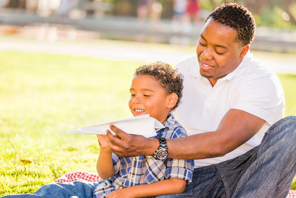 Szczęśliwy Afroamerykanin ojciec i mieszane rasy syn gry z papierowych samolotów w parku. - Zdjęcie, obraz