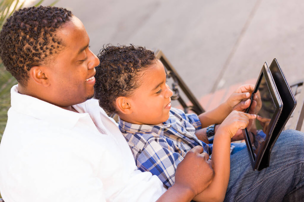 Padre afroamericano e hijo de raza mixta usando Tablet de computadora en el banco en Park. - Foto, imagen