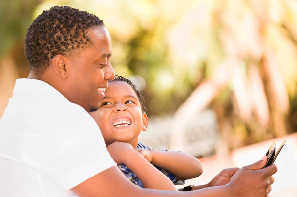 African American Father and Mixed Race Son Using Computer Tablet on Bench in Park. - 写真・画像