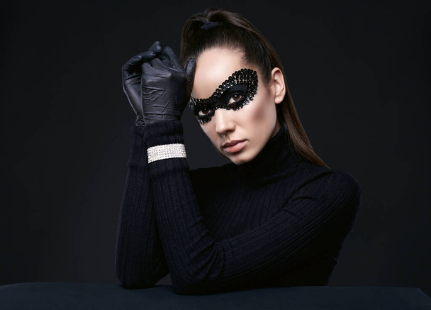 Portrait of charming elegant brunette woman in turtleneck sweater and sequins mask posing on black background in studio - 写真・画像