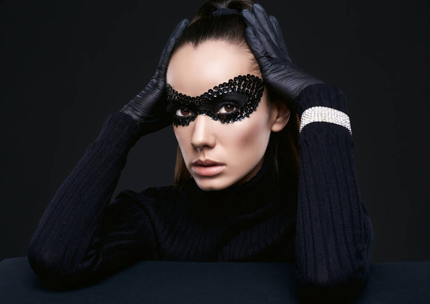 Retrato de encantadora mujer morena elegante en jersey de cuello alto y máscara de lentejuelas posando sobre fondo negro en estudio - Foto, Imagen