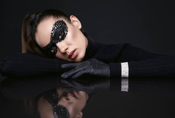 Portré bájos elegáns barna nő fekete garbó pulóver és flitterek maszk pózol egy tükrözött felületen stúdió - Fotó, kép