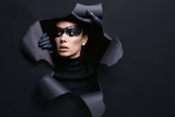 Portrait de charmante femme brune élégante en pull col roulé noir et masque à paillettes regarde à travers un fond studio déchiré - Photo, image
