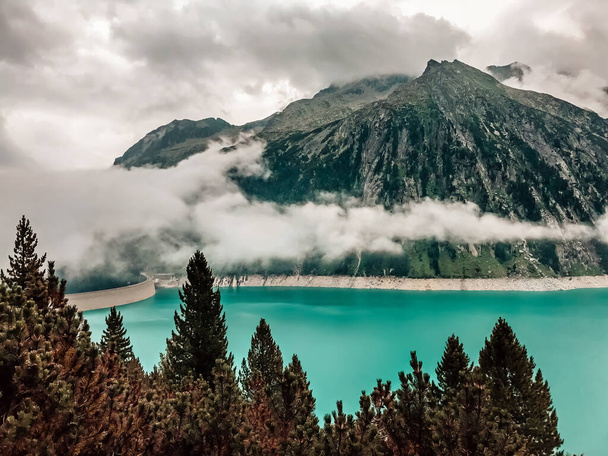 Blick durch den Fichtenwald auf das türkisfarbene Wasser des Schlegeisbeckens, einem von Nebel und Wolken verhüllten Bergreservoir. Zillertal, Mayrhofen, Österreich - Foto, Bild