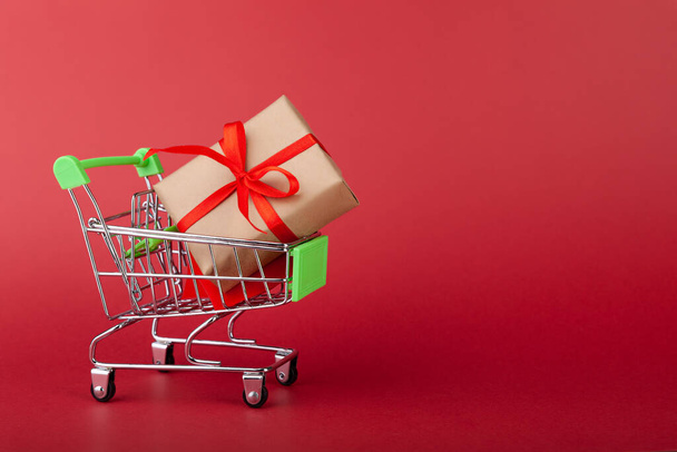 Caja de regalo con cinta roja en mini carrito de comestibles sobre fondo rosa - Foto, imagen