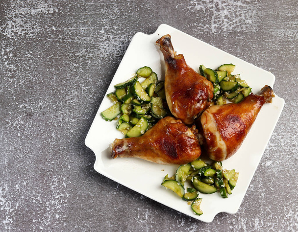 Baked chicken drumsticks with cucumber salad on a white rectangular plate on a dark background. Top view, flat lay - Fotó, kép