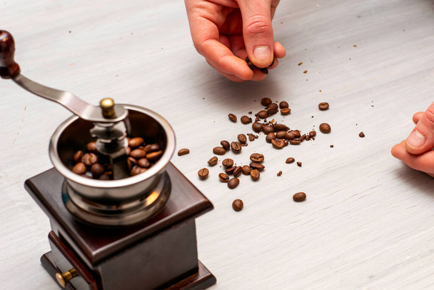 ένα άτομο αλέθει κόκκους καφέ σε ένα χειροκίνητο μύλο καφέ με τα χέρια του βάζει τους κόκκους στο μύλο καφέ χυθεί τα δημητριακά στο τραπέζι υψηλής ποιότητας φωτογραφία - Φωτογραφία, εικόνα