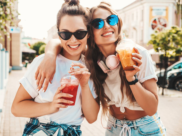 Two young beautiful smiling hipster female in trendy summer clothes. Carefree women posing outdoors.Positive models holding and drinking fresh cocktail smoothie drink in plastic cup with straw - Foto, afbeelding