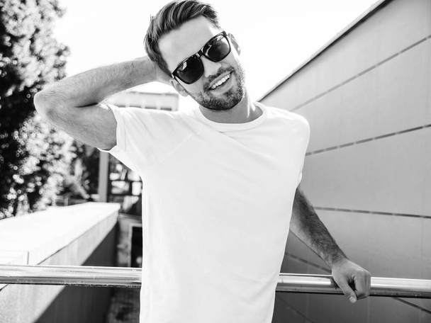 Portrait of handsome smiling stylish hipster lambersexual model.Man dressed in white T-shirt. Fashion male posing on the street background in sunglasses - Foto, Bild