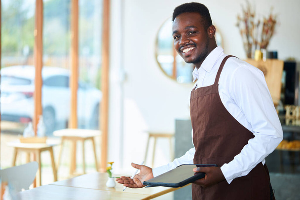 A black waiter or small restaurant operator invites customers to sit at the dining table. - Photo, Image