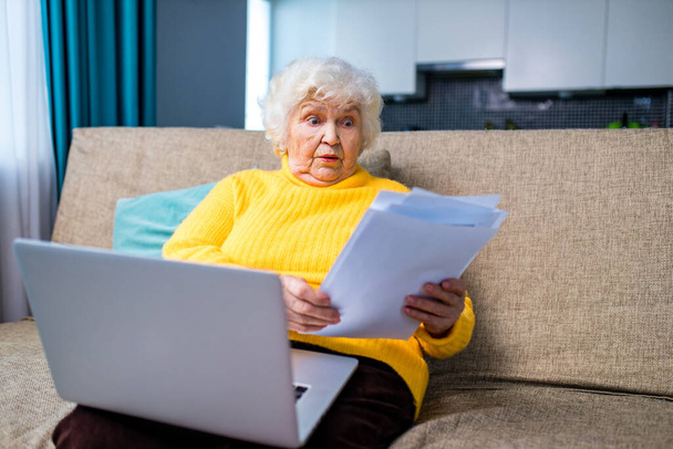shoked amazed old woman white gray-haired sitting on the sofa in living room with laptop and bills - Фото, изображение