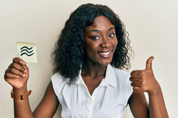 Piękna afrykańska młoda kobieta trzyma papier ze znakiem zodiaku Aquarius uśmiechając szczęśliwy i pozytywny, kciuk do góry robi doskonałe i znak aprobaty  - Zdjęcie, obraz