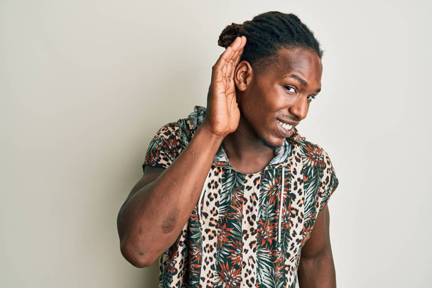 African american man with braids wearing fashion modern shirt smiling with hand over ear listening and hearing to rumor or gossip. deafness concept.  - Фото, изображение
