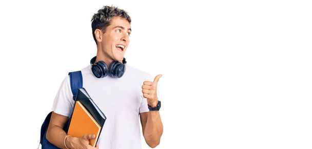 Young handsome man holding student backpack and books pointing thumb up to the side smiling happy with open mouth  - Φωτογραφία, εικόνα