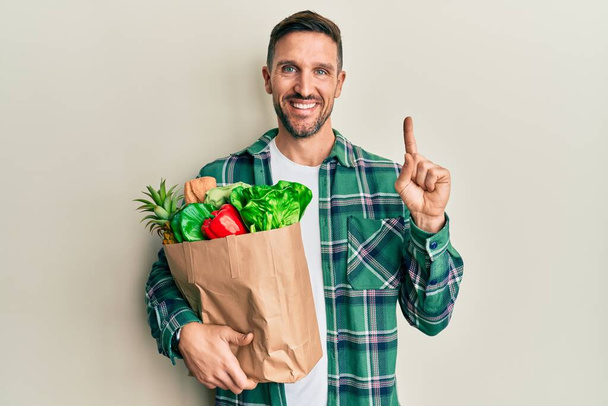 Handsome man with beard holding paper bag with groceries with a big smile on face, pointing with hand finger to the side looking at the camera.  - Photo, Image