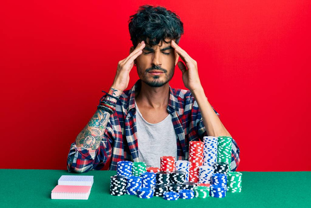 Mladý hispánec sedí na stole s pokerovými žetony a kartami s rukou na hlavě, bolest hlavy, protože stres. trpící migréna.  - Fotografie, Obrázek