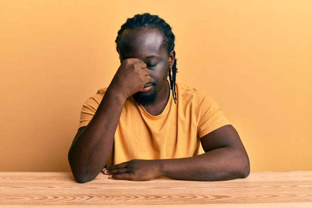 Handsome young black man wearing casual clothes sitting on the table tired rubbing nose and eyes feeling fatigue and headache. stress and frustration concept.  - Foto, imagen