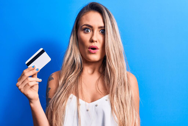 Young beautiful blonde woman holding credit card over isolated blue background scared and amazed with open mouth for surprise, disbelief face - Фото, зображення