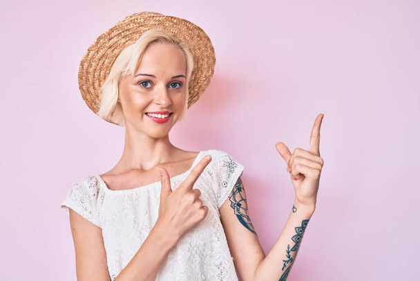 Young blonde woman with tattoo wearing summer hat smiling and looking at the camera pointing with two hands and fingers to the side.  - Foto, imagen