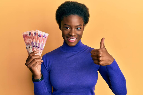 Молода афроамериканська дівчина, що тримає 50 ранд південноафриканської банкноти, усміхаючись, щаслива і позитивна, великий палець вгорі робить відмінний знак схвалення.  - Фото, зображення