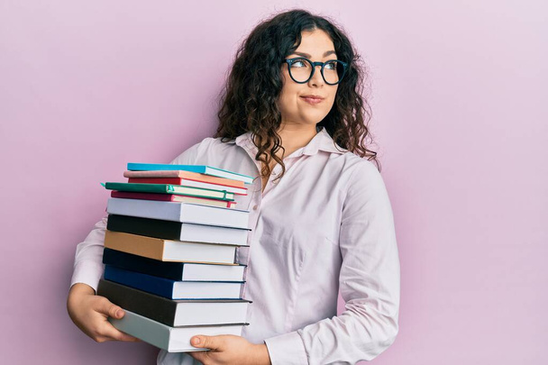 Young brunette woman with curly hair holding a pile of books smiling looking to the side and staring away thinking.  - Photo, Image