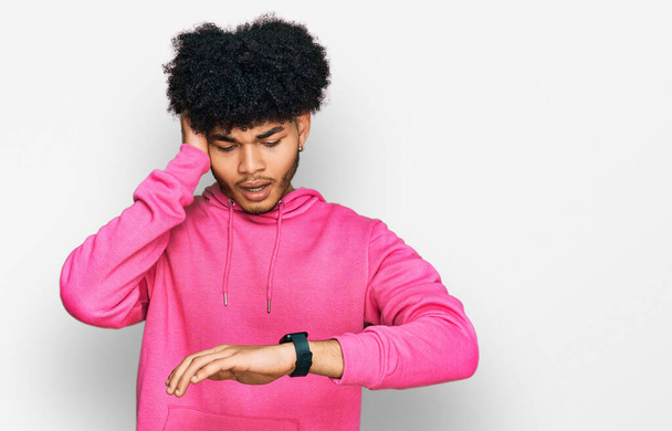Junger afrikanisch-amerikanischer Mann mit Afro-Haaren trägt lässiges rosafarbenes Sweatshirt und schaut besorgt auf die Uhrzeit, Angst, zu spät zu kommen  - Foto, Bild
