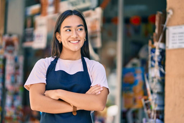 jong latin winkelier meisje met armen gekruist lachend gelukkig in de pers winkel - Foto, afbeelding