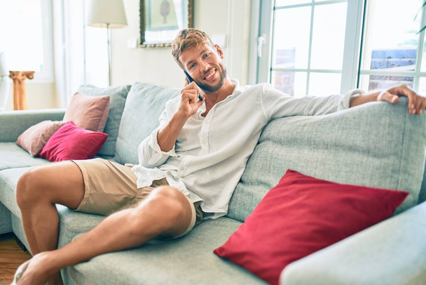 Yakışıklı beyaz adam gülümsüyor. Evdeki kanepede oturup telefonla konuşuyor. - Fotoğraf, Görsel