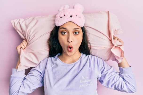 Beautiful middle eastern woman wearing sleep mask and pajama sleeping on pillow afraid and shocked with surprise and amazed expression, fear and excited face.  - Photo, Image