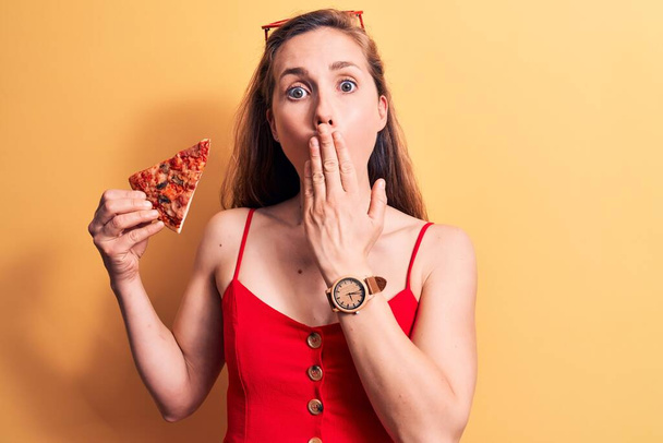 Joven mujer rubia hermosa comiendo una rebanada de pizza con vestido de verano que cubre la boca con la mano, conmocionada y asustada por el error. expresión sorprendida  - Foto, Imagen