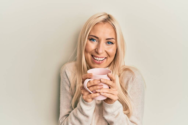 Young blonde woman drinking a cup of coffee smiling and laughing hard out loud because funny crazy joke.  - Fotografie, Obrázek