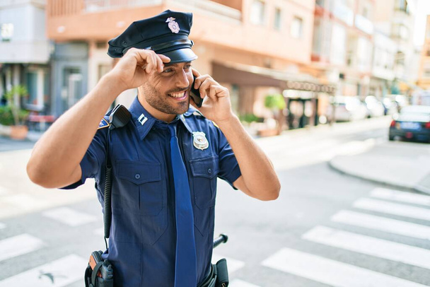 A fiatal jóképű spanyol rendőr, aki rendőregyenruhát visel, boldogan mosolyog. Álló mosollyal az arcán beszélgetnek az okostelefonon a város utcájában. - Fotó, kép