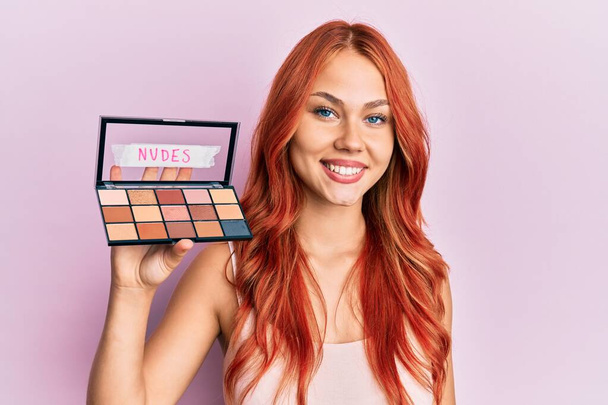 Young redhead woman holding makeup nudes looking positive and happy standing and smiling with a confident smile showing teeth  - Photo, Image