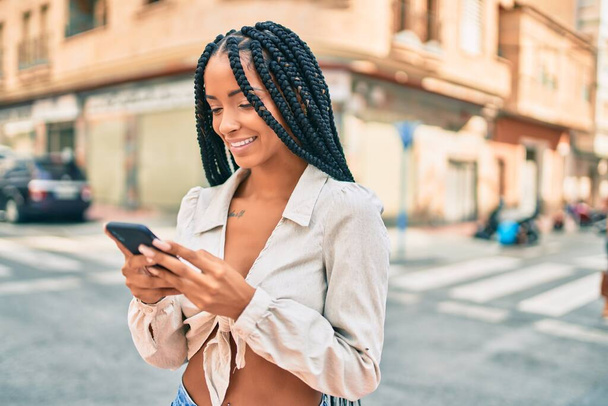 Νεαρή Αφροαμερικανή γυναίκα χαμογελά ευτυχισμένη χρησιμοποιώντας smartphone στην πόλη. - Φωτογραφία, εικόνα