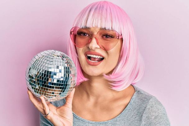 Joven mujer caucásica vistiendo peluca rosa sosteniendo bola disco mirando positiva y feliz de pie y sonriendo con una sonrisa confiada mostrando los dientes  - Foto, Imagen