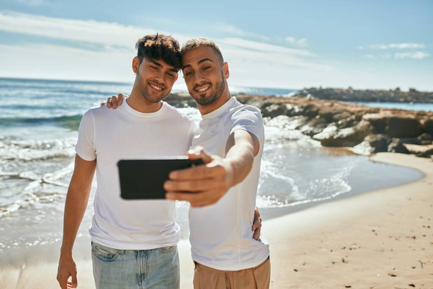 Νεαρό γκέι ζευγάρι χαμογελά χαρούμενος κάνοντας selfie από το smartphone στην παραλία. - Φωτογραφία, εικόνα