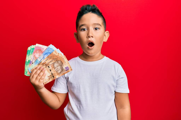 Niño hispano sosteniendo billetes de rand sudafricanos asustado y sorprendido con la boca abierta para sorpresa, cara de incredulidad  - Foto, Imagen