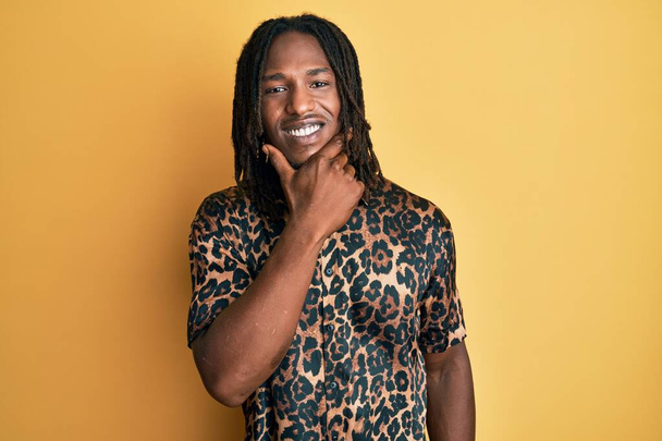 Homem afro-americano com tranças vestindo camisa de impressão animal leopardo olhando confiante para a câmera sorrindo com os braços cruzados e mão levantada no queixo. pensamento positivo.  - Foto, Imagem